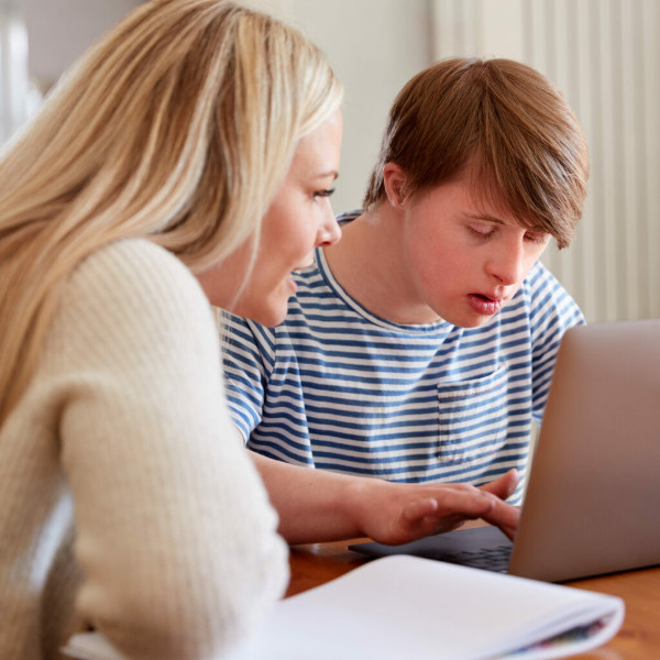 Downs Syndrome Man Sitting With Home Tutor Using Laptop For Lesson At Home